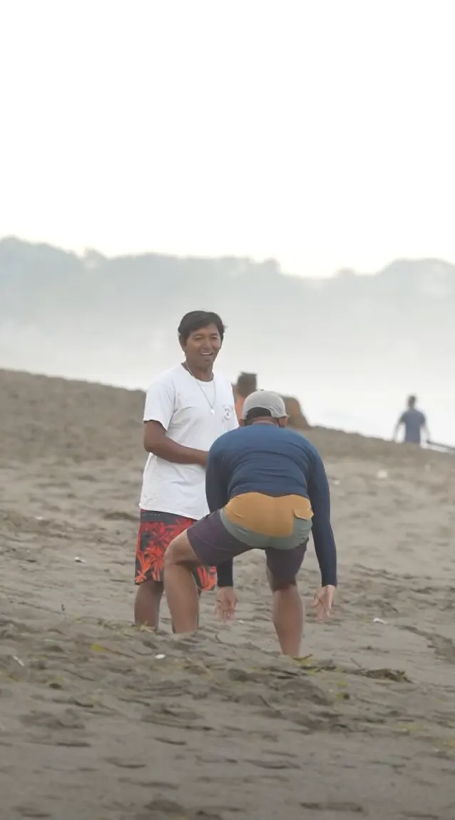 Two surfers share a laugh on the beach, one crouched playfully in the sand