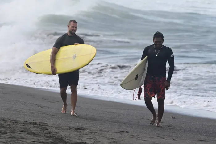 Two surfers carrying their boards walk up the beach as a wave breaks behind them